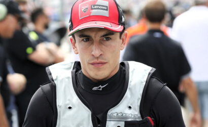 Male motorcycle racer in a red Lenovo cap and light protective vest, standing in a busy pit lane with a focused expression.