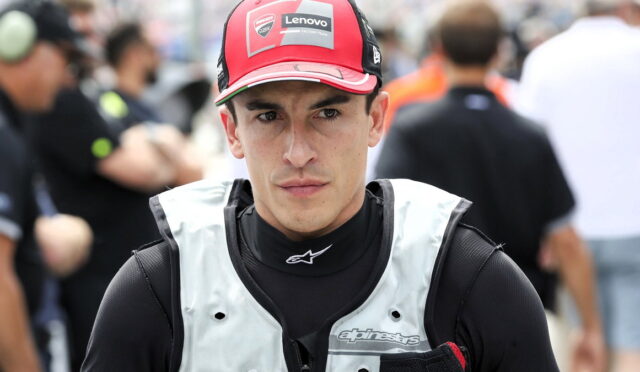 Male motorcycle racer in a red Lenovo cap and light protective vest, standing in a busy pit lane with a focused expression.