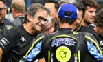 Smiling man with glasses talks to a motorcycle racer in a blue cap, surrounded by team members in a busy paddock.