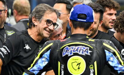 Smiling man with glasses talks to a motorcycle racer in a blue cap, surrounded by team members in a busy paddock.