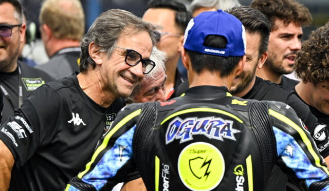 Smiling man with glasses talks to a motorcycle racer in a blue cap, surrounded by team members in a busy paddock.