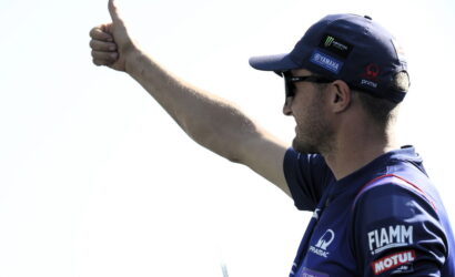 Side view of a man wearing a blue cap and sunglasses, giving a thumbs up with his right arm visible and sponsor logos on his sleeve.