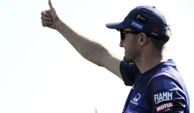 Side view of a man wearing a blue cap and sunglasses, giving a thumbs up with his right arm visible and sponsor logos on his sleeve.