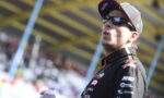 Motorsport racer wearing a black cap and sunglasses, holding a water bottle, on a racetrack with a blurred crowd behind him.