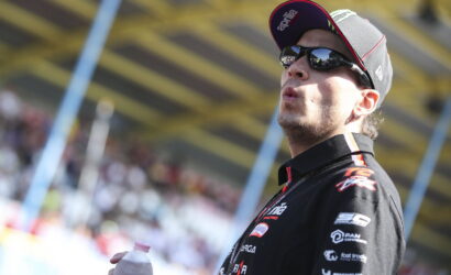 Motorsport racer wearing a black cap and sunglasses, holding a water bottle, on a racetrack with a blurred crowd behind him.