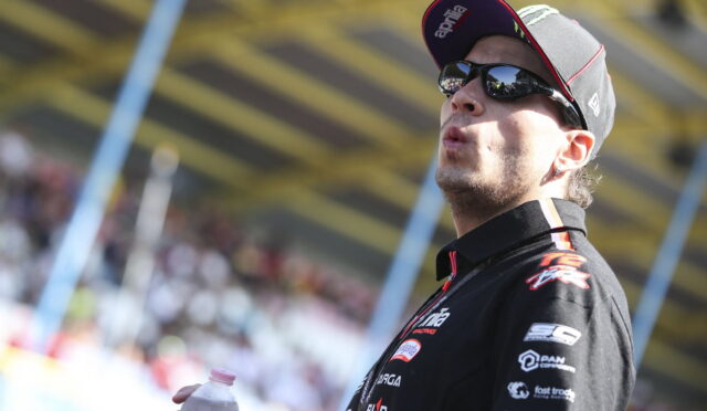 Motorsport racer wearing a black cap and sunglasses, holding a water bottle, on a racetrack with a blurred crowd behind him.