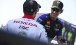 Two motorcycle racers chat and smile in a sunny paddock; one in a blue Yamaha jersey and Monster cap.