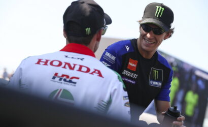 Two motorcycle racers chat and smile in a sunny paddock; one in a blue Yamaha jersey and Monster cap.