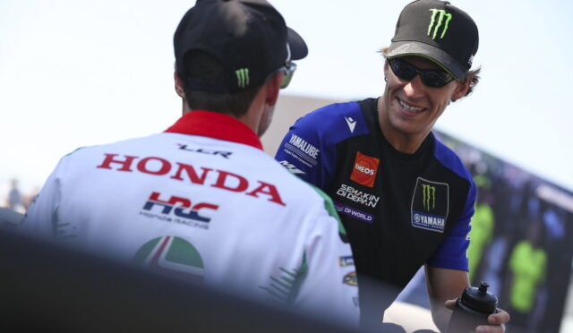 Two motorsport team members converse in a paddock; one wearing a white Honda Racing jacket, the other in a Yamaha cap.