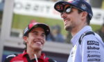 Two race team members smiling and chatting at a trackside event; man in a blue cap and sunglasses alongside another in red gear.