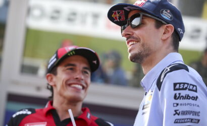 Two race team members smiling and chatting at a trackside event; man in a blue cap and sunglasses alongside another in red gear.