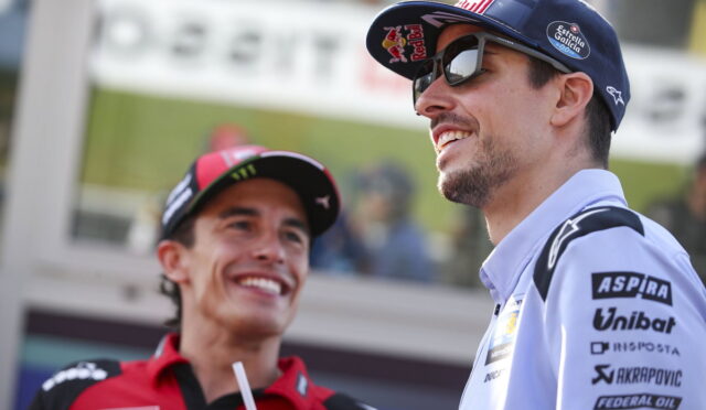 Two race team members smiling and chatting at a trackside event; man in a blue cap and sunglasses alongside another in red gear.