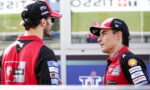Two race team members in red and black uniforms talk in a pit lane area, facing each other.