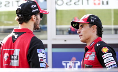 Two race team members in red and black uniforms talk in a pit lane area, facing each other.