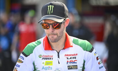 Male motorcycle racer in white, green, and red Honda racing jersey wearing a black Monster cap and orange mirrored sunglasses with a water bottle in hand.