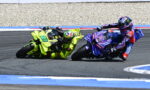 Two motorcycle racers lean into a left-hand turn on a race track, one bike in neon yellow and the other in blue/purple livery.