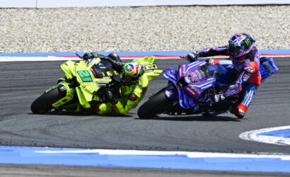 Two motorcycle racers lean into a left-hand turn on a race track, one bike in neon yellow and the other in blue/purple livery.