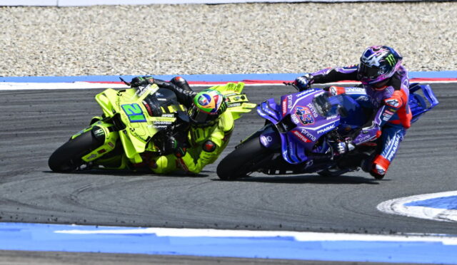 Two motorcycle racers lean into a left-hand turn on a race track, one bike in neon yellow and the other in blue/purple livery.