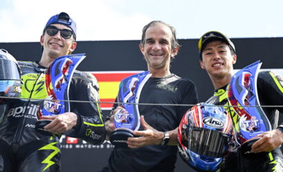 Three motorcyclists stand together on a podium, each holding a blue trophy and smiling at the camera.
