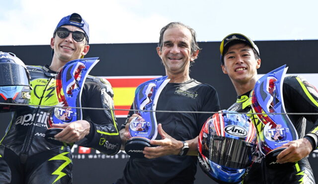 Three motorcyclists stand together on a podium, each holding a blue trophy and smiling at the camera.