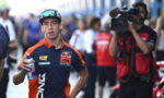 Motorsport rider in Red Bull KTM gear holding a water bottle, with a camera crew in the background in a busy pit area or paddock.