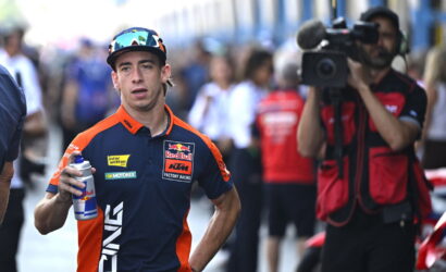 Motorsport rider in Red Bull KTM gear holding a water bottle, with a camera crew in the background in a busy pit area or paddock.