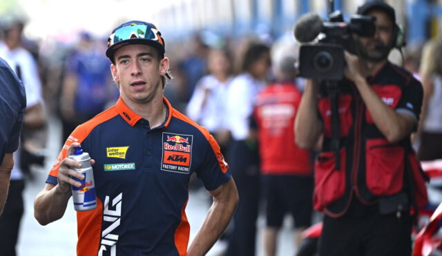 Motorsport rider in Red Bull KTM gear holding a water bottle, with a camera crew in the background in a busy pit area or paddock.