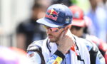 Male motorbike racer wearing a blue Red Bull cap and mirrored sunglasses, adjusting his ear while in a racing suit with a towel over his shoulders.