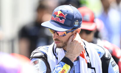 Male motorbike racer wearing a blue Red Bull cap and mirrored sunglasses, adjusting his ear while in a racing suit with a towel over his shoulders.