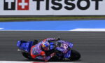 MotoGP rider on a blue racing motorcycle leans into a turn on a race track, with a Tissot banner in the background and red accents on the rider's suit and bike.