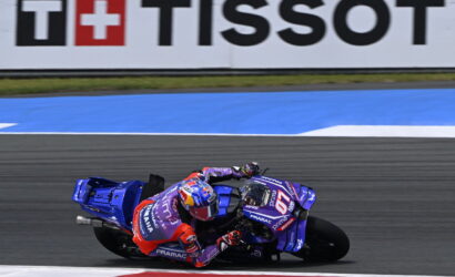 MotoGP rider on a blue racing motorcycle leans into a turn on a race track, with a Tissot banner in the background and red accents on the rider's suit and bike.