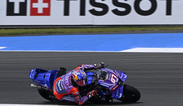 MotoGP rider on a blue racing motorcycle leans into a turn on a race track, with a Tissot banner in the background and red accents on the rider's suit and bike.