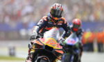 MotoGP rider in a Red Bull KTM racing suit on an orange racing bike, leaning into a turn on the track with a blurred crowd background