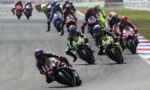 Motorcycle race with riders leaning into a curve, leading rider wearing red/black ahead of a pack on a track surface with red-white-blue run-off borders.