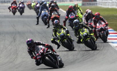 Motorcycle race with riders leaning into a curve, leading rider wearing red/black ahead of a pack on a track surface with red-white-blue run-off borders.