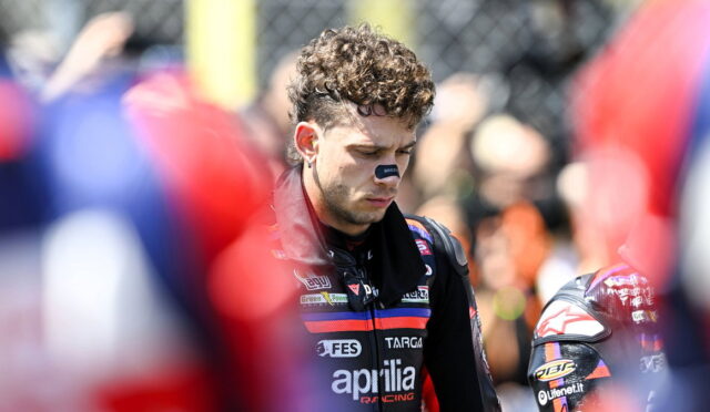 Male motorcycle racer in an Aprilia Racing suit with eye black, looking down in a pit area.