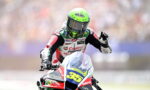 Motorsport rider in a neon green helmet on a red, white, and blue race bike with number 65, waving to the crowd.