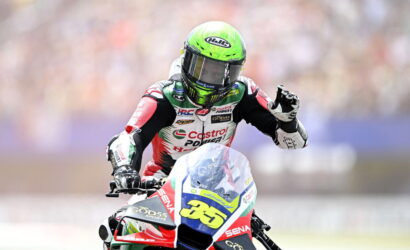 Motorsport rider in a neon green helmet on a red, white, and blue race bike with number 65, waving to the crowd.