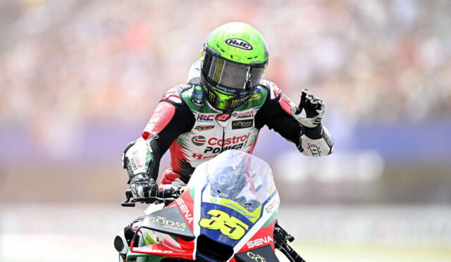 Motorsport rider in a neon green helmet on a red, white, and blue race bike with number 65, waving to the crowd.