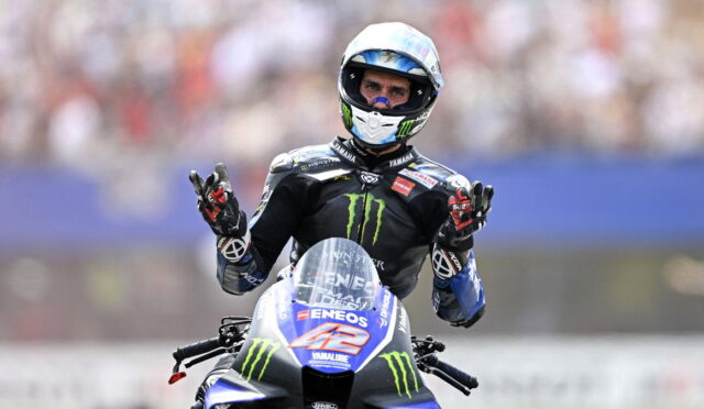 MotoGP rider in a black Monster Yamaha suit on a Yamaha bike, hands raised in celebration.
