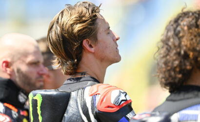 Male motorcycle racer in protective gear, side profile with eyes closed, at a track event; Monster Energy logo on shoulder pad visible