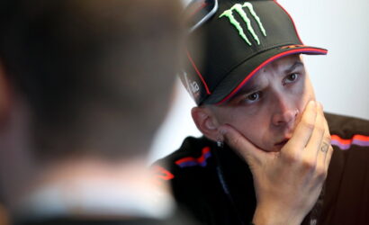 Man wearing a black Monster cap with sunglasses perched on top, resting his chin on his hand in thought.