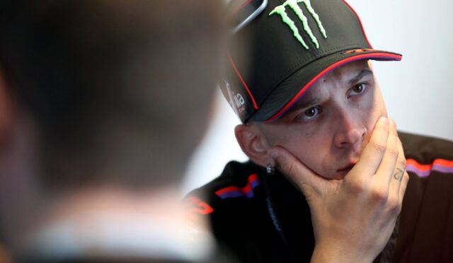 Man wearing a black Monster cap with sunglasses perched on top, resting his chin on his hand in thought.