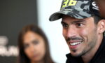 Close-up of a smiling man wearing a camouflage cap, with a blurred woman in the background.