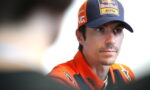 Portrait of a man in an orange KTM racing jacket and a Red Bull cap, looking to the side in an interview setting.