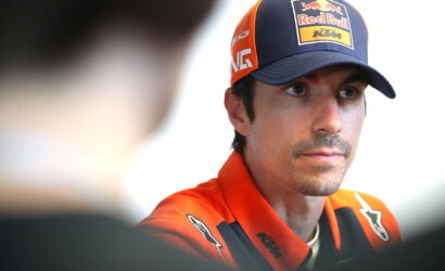 Portrait of a man in an orange KTM racing jacket and a Red Bull cap, looking to the side in an interview setting.