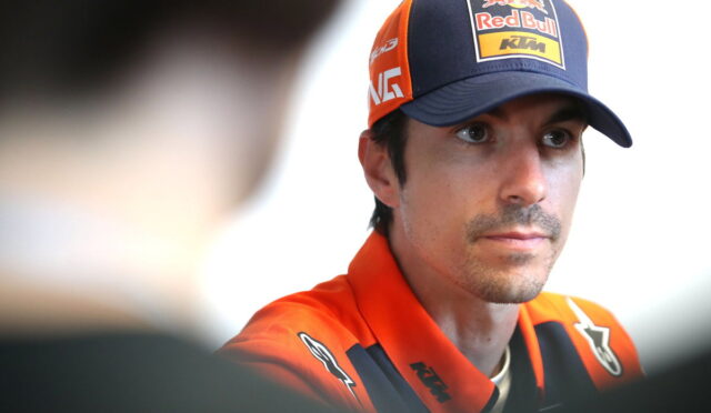 Portrait of a man in an orange KTM racing jacket and a Red Bull cap, looking to the side in an interview setting.