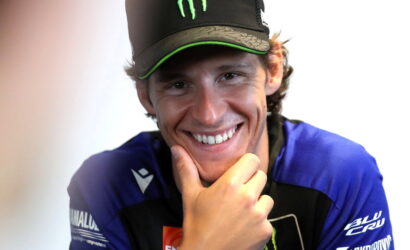 Young man in a blue racing jersey and black cap with a green Monster logo, smiling and resting his chin on his hand.