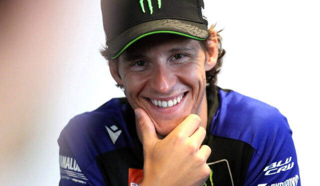 Young man in a blue racing jersey and black cap with a green Monster logo, smiling and resting his chin on his hand.