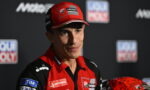 Portrait of a male MotoGP rider in a red and black Ducati Lenovo racing suit at a press event, wearing a red cap with sponsor logos and a red helmet nearby.